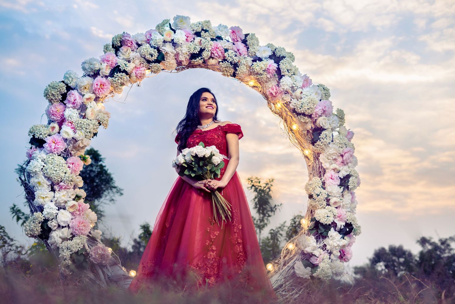 A figure in a flowing red gown poses beneath a romantic floral arch of pink and white blooms at sunset.