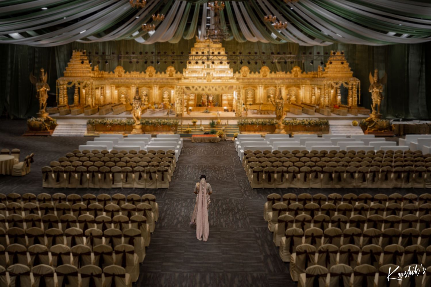 An elegant wedding ceremony setup with gold decor and draped fabric in a grand ballroom with rows of wooden chairs.