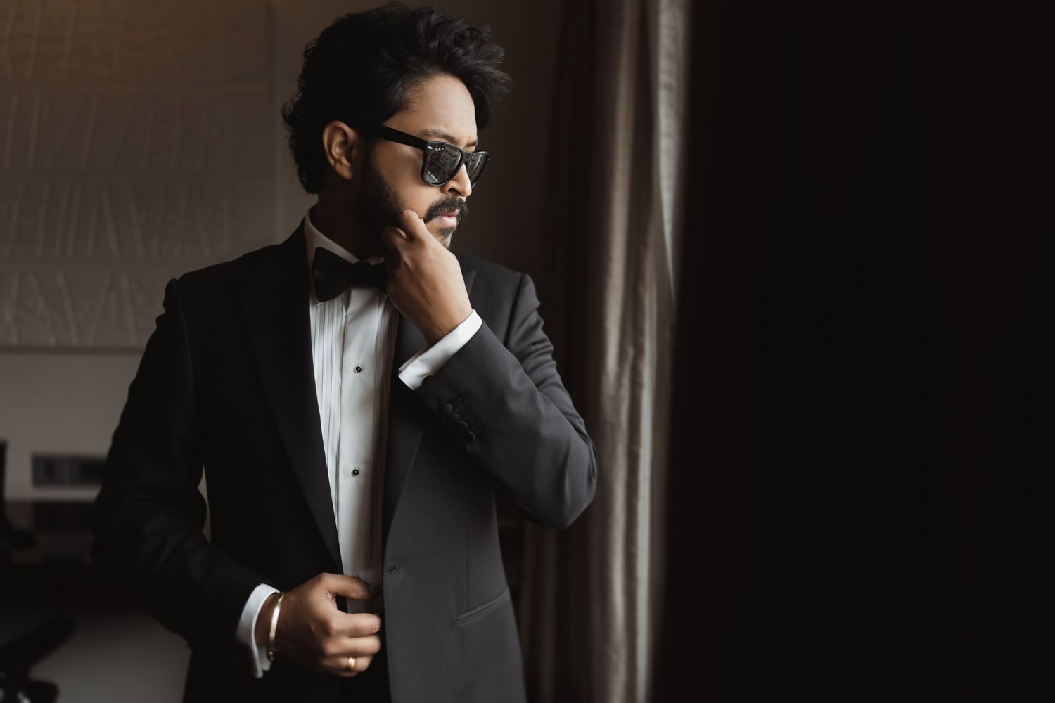 Stylishly dressed figure in black tuxedo and bow tie stands thoughtfully by window with hand near face.