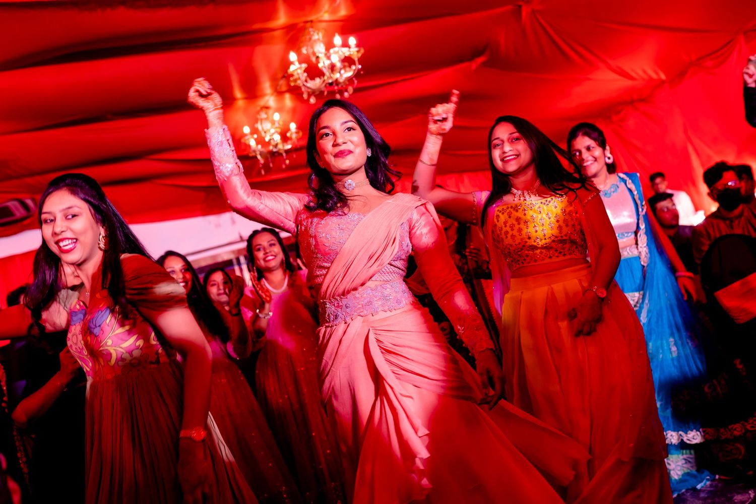 People in colorful traditional attire dancing at a festive celebration bathed in vibrant red lighting.