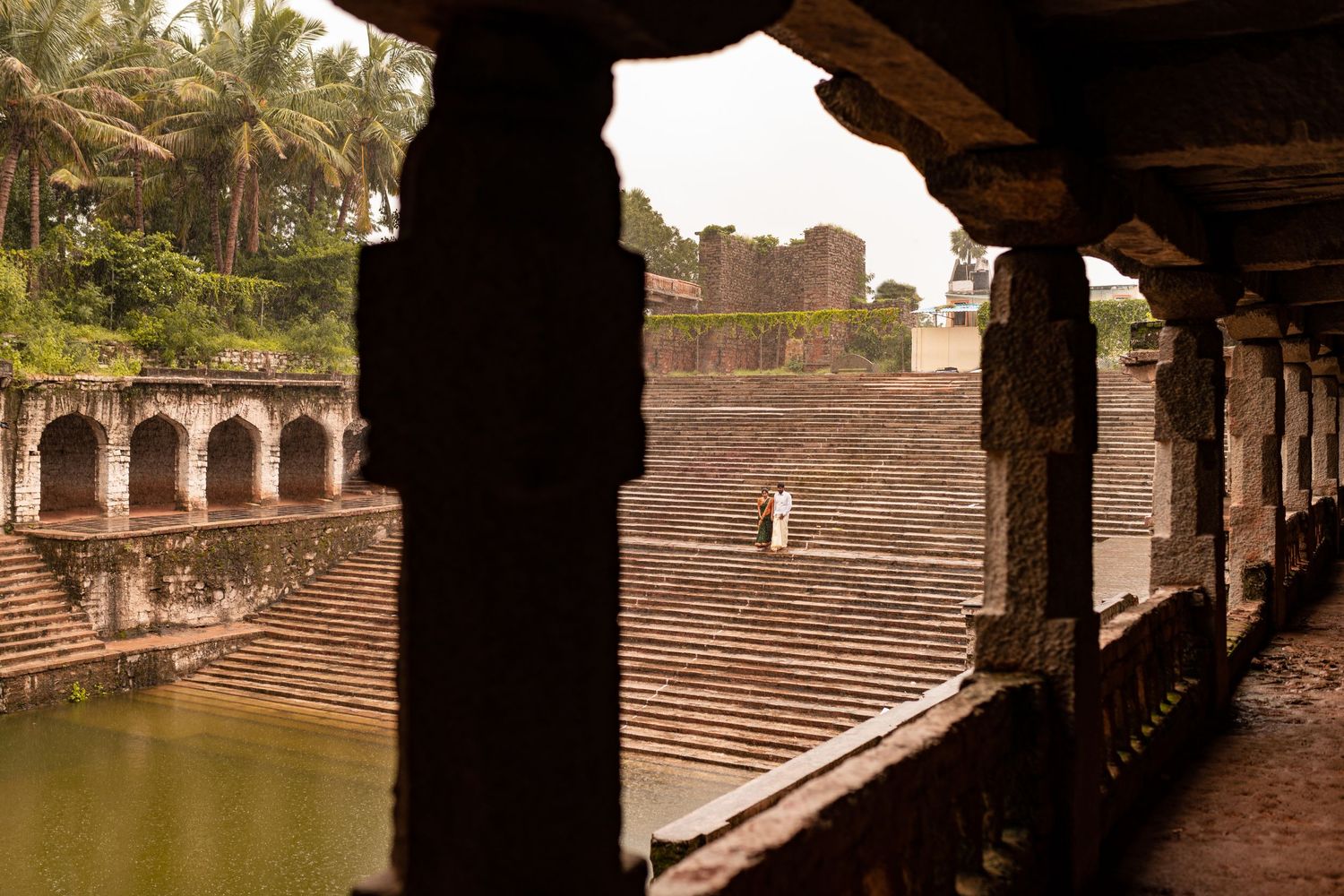 Ancient stone steps and archways overlook a historic water reservoir at a temple complex with palm trees in the background.