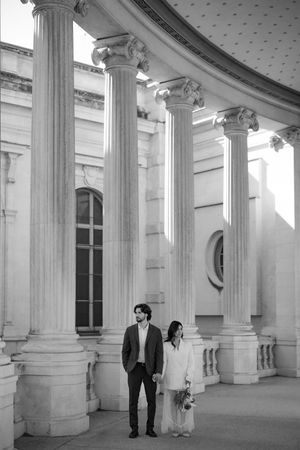 Black and white photo of a couple holding hands among grand columns of a classical building.
