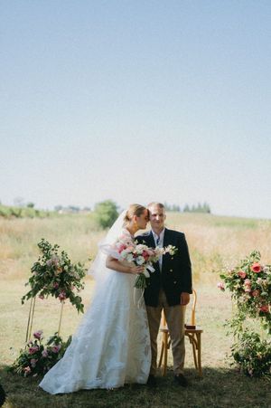 Bride in lace gown holding pink floral bouquet kisses man in navy blazer at outdoor wedding ceremony.