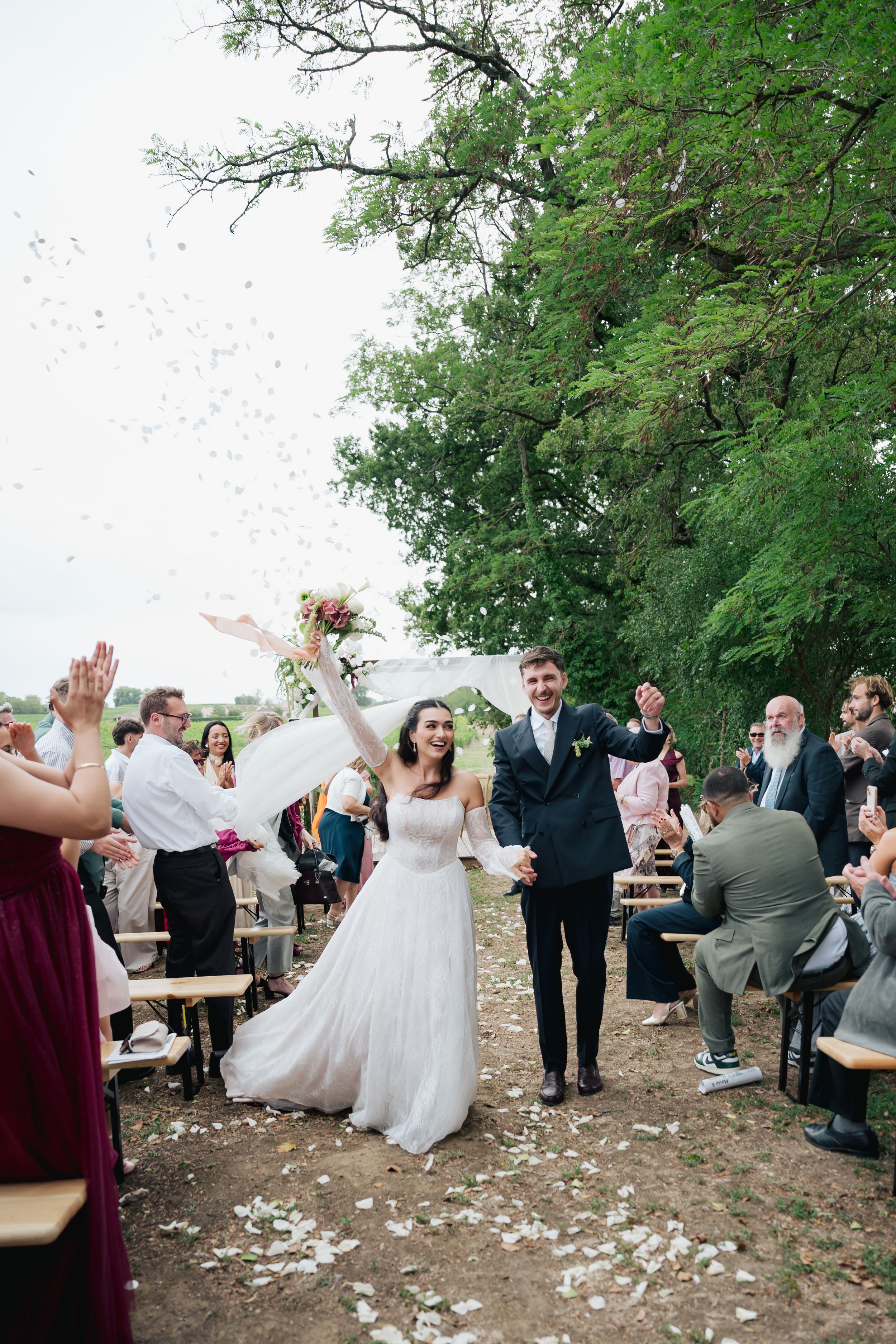 Joyful newlyweds walk down outdoor aisle as guests toss white flower petals during sunny wedding ceremony.