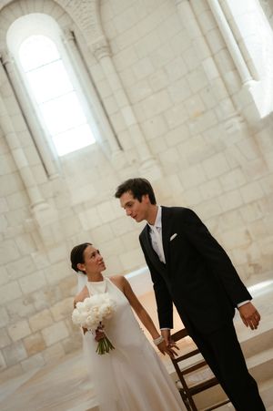 Bride holding white bouquet and groom in black suit share a tender moment inside a stone church.
