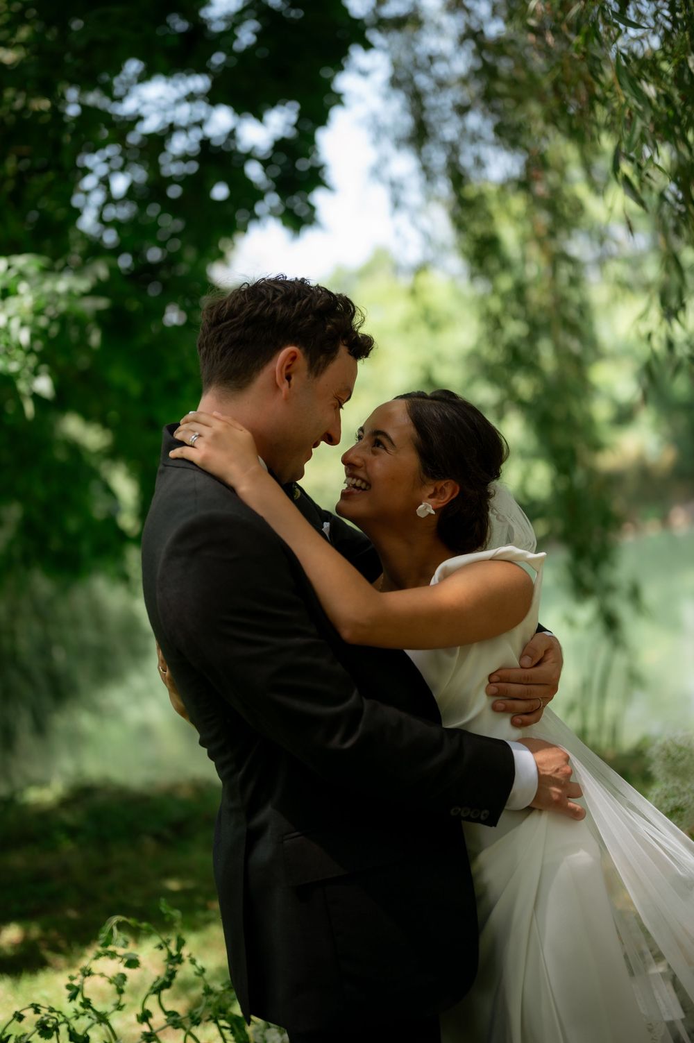 Bride and groom embrace under lush green trees, smiling at each other in an romantic outdoor wedding portrait.