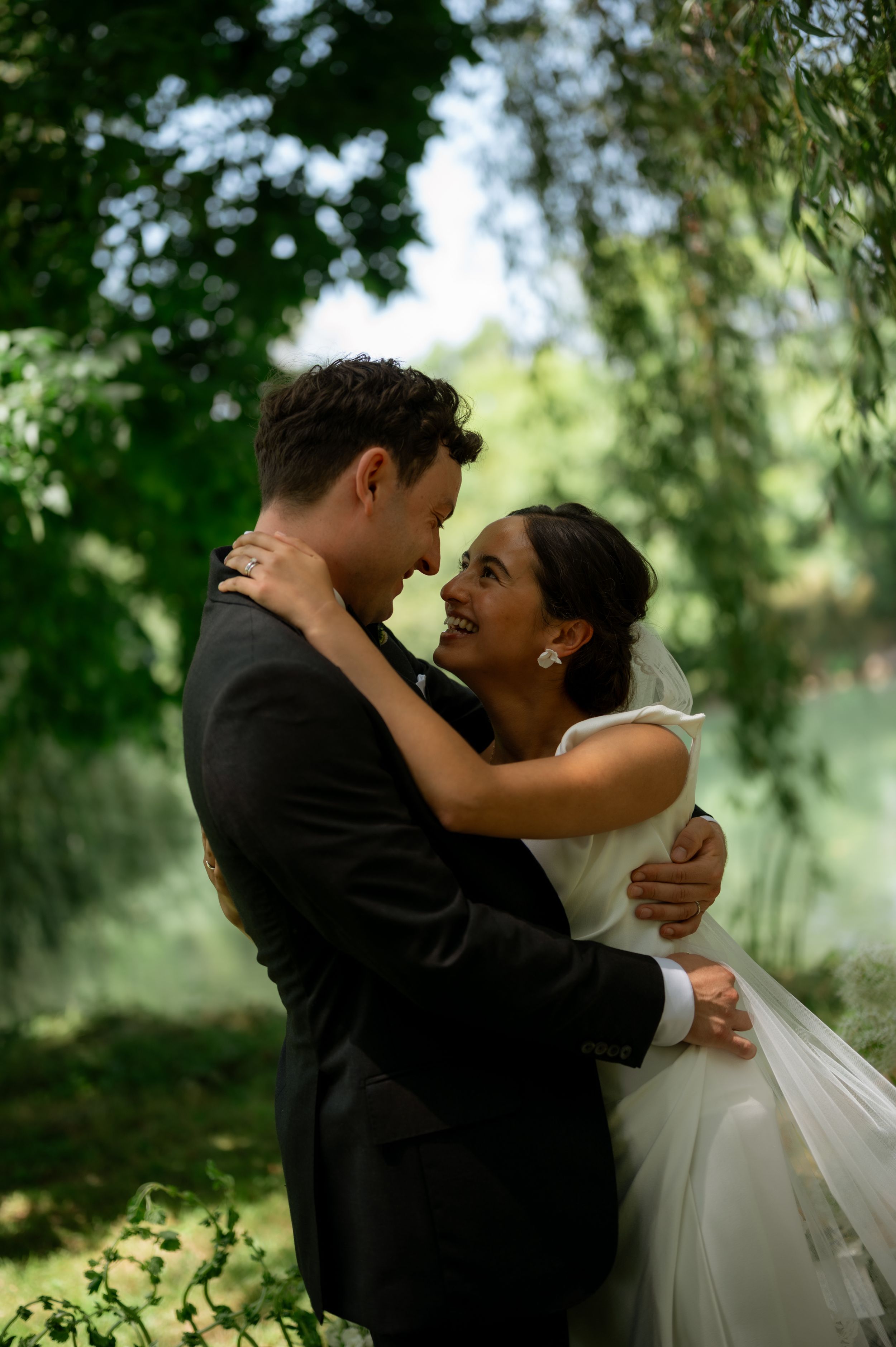 Bride and groom embrace under lush green trees, smiling at each other in an romantic outdoor wedding portrait.