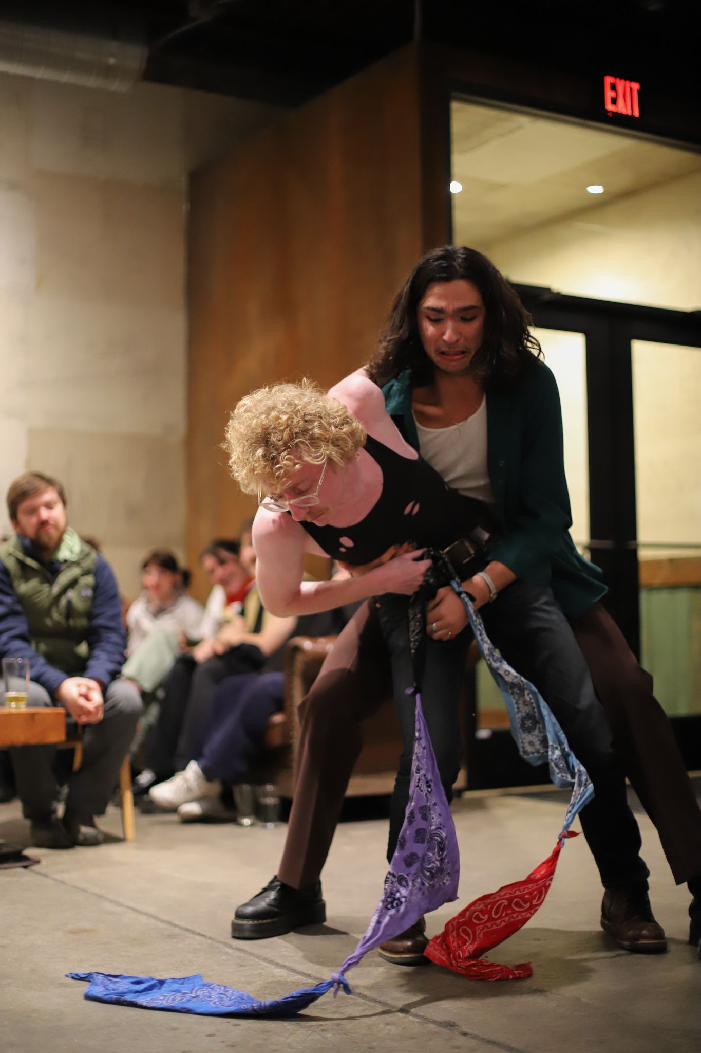 Two performers on stage doing physical theater with colorful fabric props while audience watches in background.