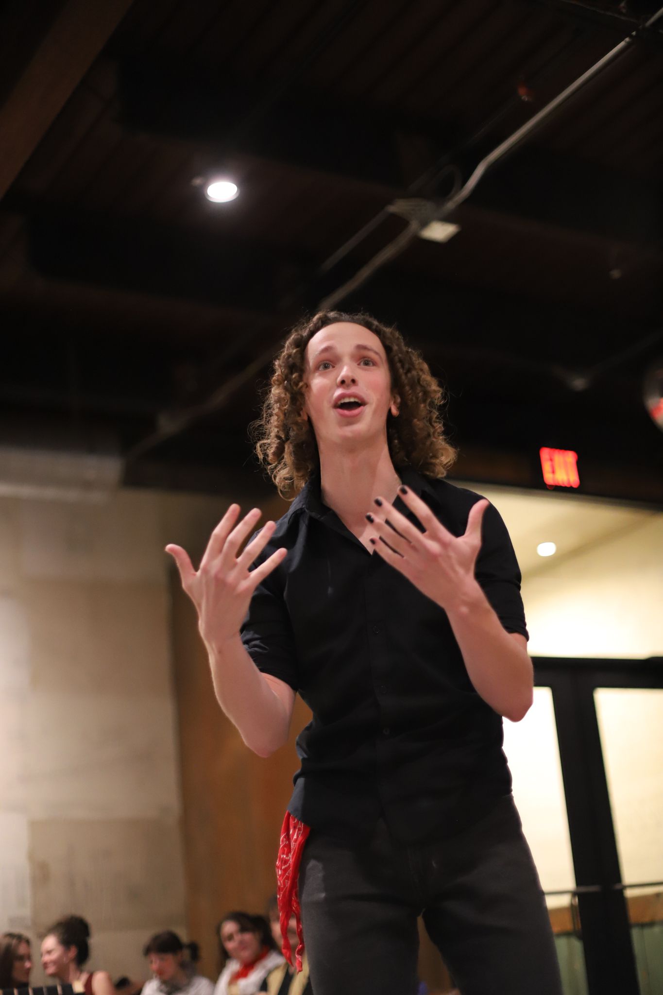 A performer in black rehearses on stage with dramatic hand gestures under dim lighting.