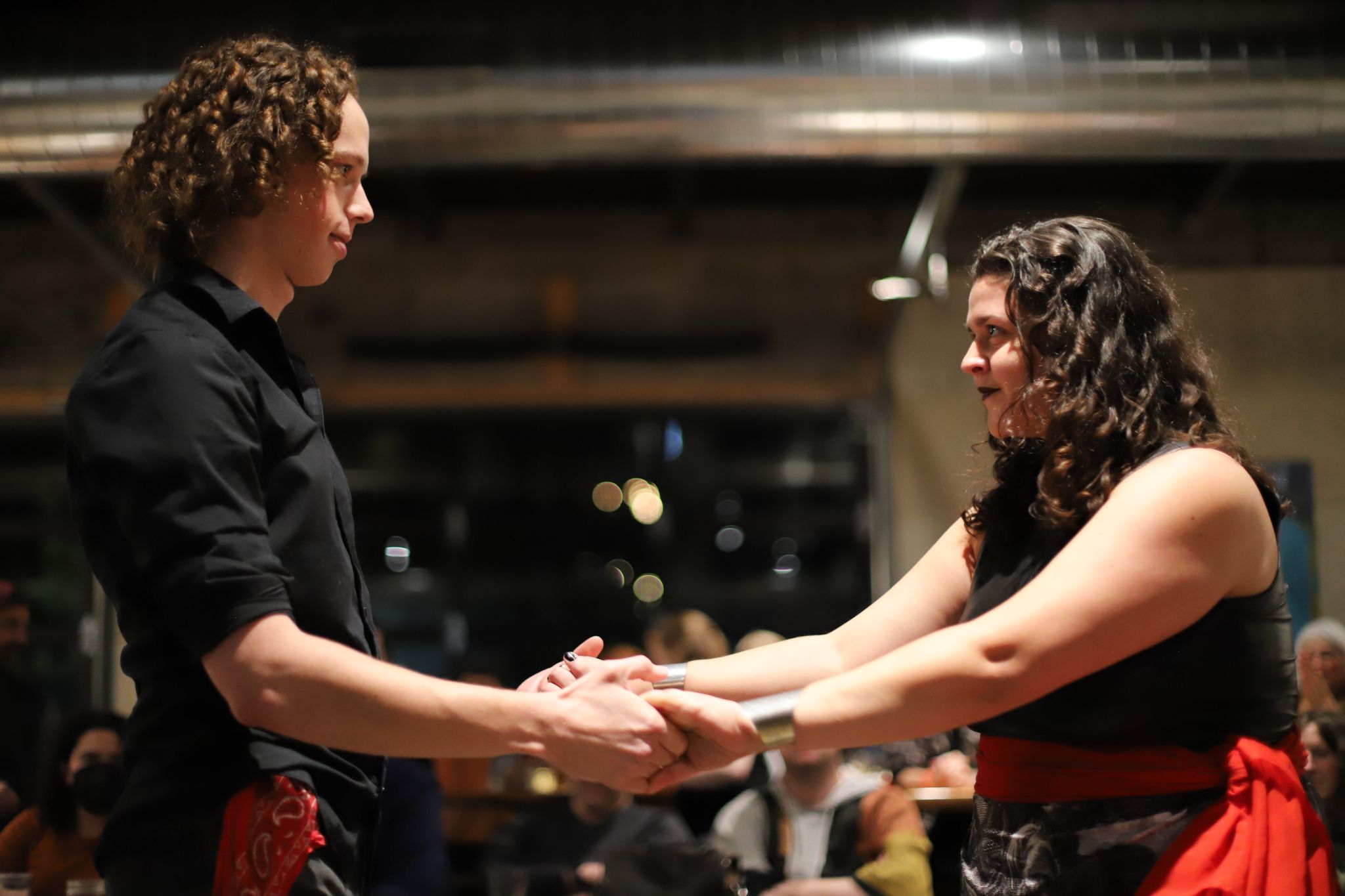 Two dancers holding hands during a performance, one wearing black and one wearing a red sash.