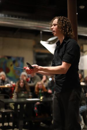A theater director wearing black conducts rehearsal with expressive gestures in front of an audience.