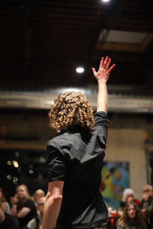 A performer with curly hair raises their hand during a performance in a dimly lit indoor venue.