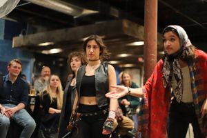 People gathered in a dimly lit indoor space with industrial ceiling, wearing casual clothing and accessories.
