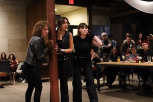 Three performers huddle together during a theatrical performance while audience members watch in background.