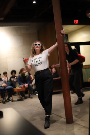 A person in sunglasses and 'I'm a Artist' t-shirt poses confidently against a pillar while holding a drink at an indoor event.