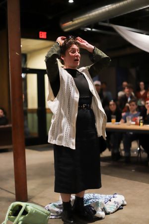 A performer in a white cardigan and black dress gestures dramatically on stage during a theatrical presentation.