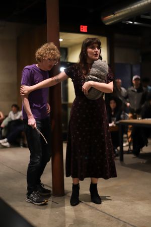 Two performers in contrasting outfits stand near a support beam during a theatrical scene.