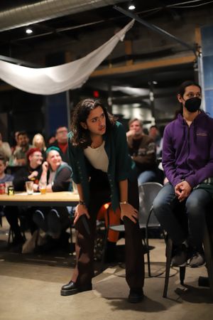 Person in dark jacket and boots leans forward while watching intently in a dimly lit room with audience members.