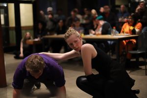 Two performers in dark clothing engage in a physical performance piece on a floor.