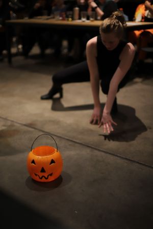 A performer in black reaches toward an orange Halloween pumpkin bucket on a dark floor.