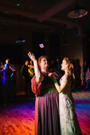 Two dancers in elegant dresses share a joyful moment under colorful party lights on a wooden dance floor.