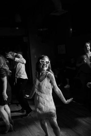 A person in a flapper-style dress and heart-shaped glasses dances joyfully at a wedding reception.
