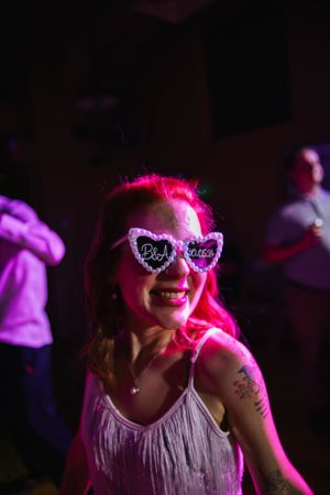 Person with bright pink hair wearing heart-shaped sunglasses and white fringe top in purple nightclub lighting.