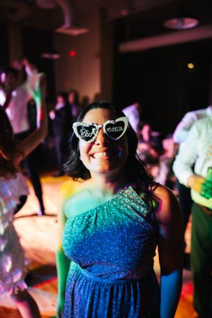 A party guest in a blue dress and heart-shaped sunglasses smiles at a celebratory event.