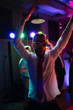 A party-goer dances with arms raised under colorful disco lights wearing a white shirt and glasses.