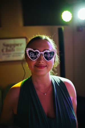 A partygoer wearing heart-shaped sunglasses smiles under colorful venue lighting.