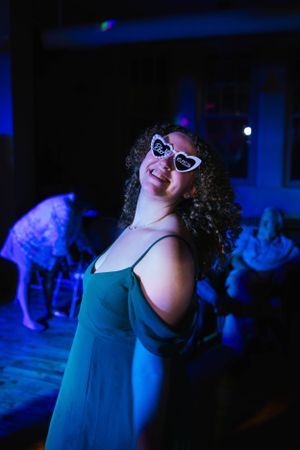A person in a teal dress and retro sunglasses poses playfully under blue lighting at a party.