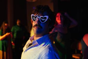 Person wearing heart-shaped sunglasses illuminated by colorful party lights against a dark background.
