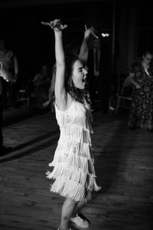 A dancer in a white fringe flapper dress performs energetically with raised arms on a wooden dance floor.