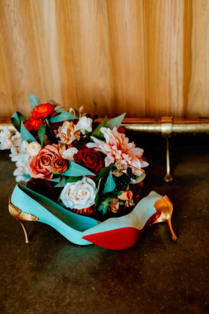 A bridal bouquet with red and pink roses sits nestled in turquoise wedding shoes against a wooden backdrop.