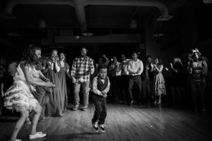 Young dancer performs on wooden dance floor during wedding reception while guests watch and enjoy in black and white photo.