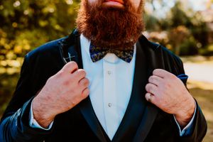 Close up of a red bearded person in formal attire adjusting a bow tie and black suit jacket.