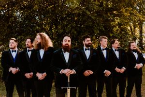 A line of groomsmen in black tuxedos and bowties smile together outdoors with autumn trees in the background.