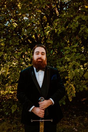 A groom in a black suit and bow tie stands with a sword against a backdrop of autumn foliage at golden hour.