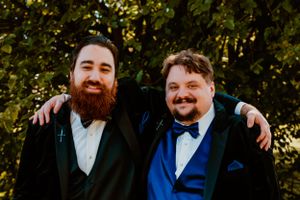 Two men in formal suits stand together outdoors, one wearing black with a beard and one in bright blue with a bow tie.