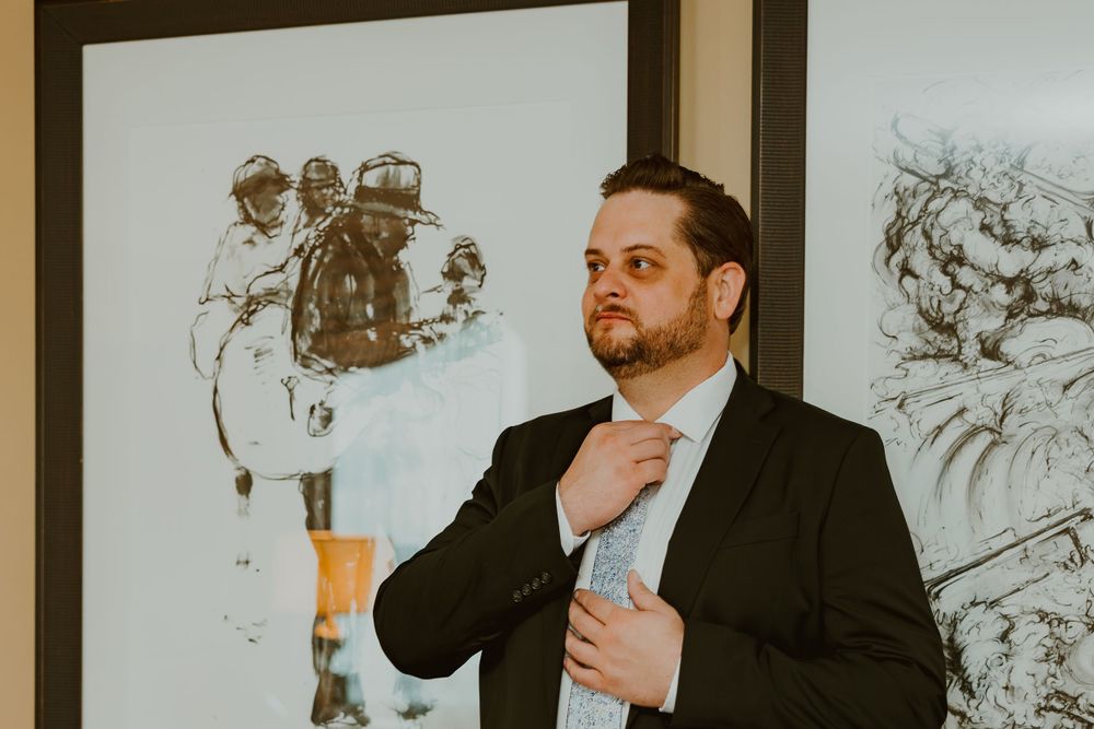 A person in a dark suit adjusts their tie while standing in front of artwork hanging on a wall.