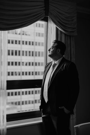 A figure in a business suit gazes contemplatively out of a high-rise window with dramatic lighting and curtains.