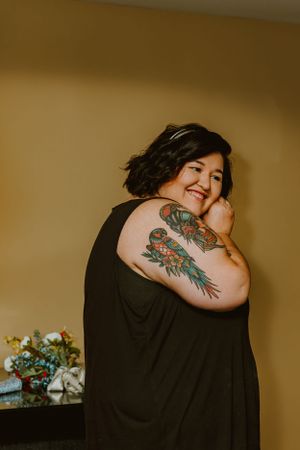 Smiling person in black dress showing colorful floral arm tattoo against warm beige wall with decorative flowers.