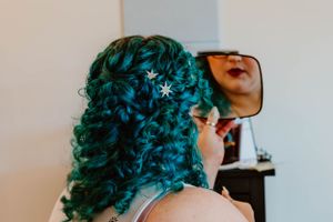 Person with vibrant turquoise curly hair adorned with small star decorations looking into a handheld mirror.