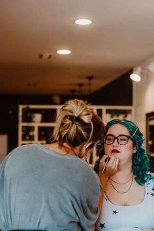 Makeup artist applies lipstick to client with bright teal hair and glasses in indoor setting.