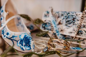 An elegant white ballet flat shoe with blue floral embroidery pattern rests among scattered leaves and fabric.