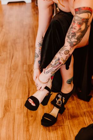 Detailed view of tattooed legs wearing black strappy sandals on wooden floor.