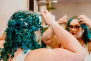 A person with vibrant turquoise curly hair adorned with a small starfish accessory is shown in a reflection.