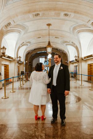 A couple stands in a grand marble hallway with decorative arches and gold accents.