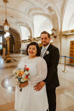 A wedding couple posing in an ornate historic building with grand arches and chandeliers.