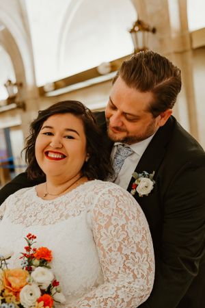 A romantic wedding photo showing a couple embracing, with the bride in lace and holding a colorful autumn bouquet.